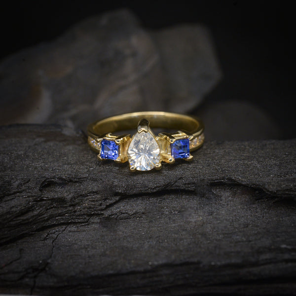Anillo de compromiso con diamante natural corte gota de .50ct, 12 moissanitas y 2 zafiros naturales corte princesa elaborado en oro amarillo de 14 kilates