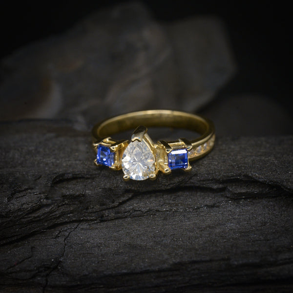 Anillo de compromiso con diamante natural corte gota de .50ct, 12 moissanitas y 2 zafiros naturales corte princesa elaborado en oro amarillo de 14 kilates